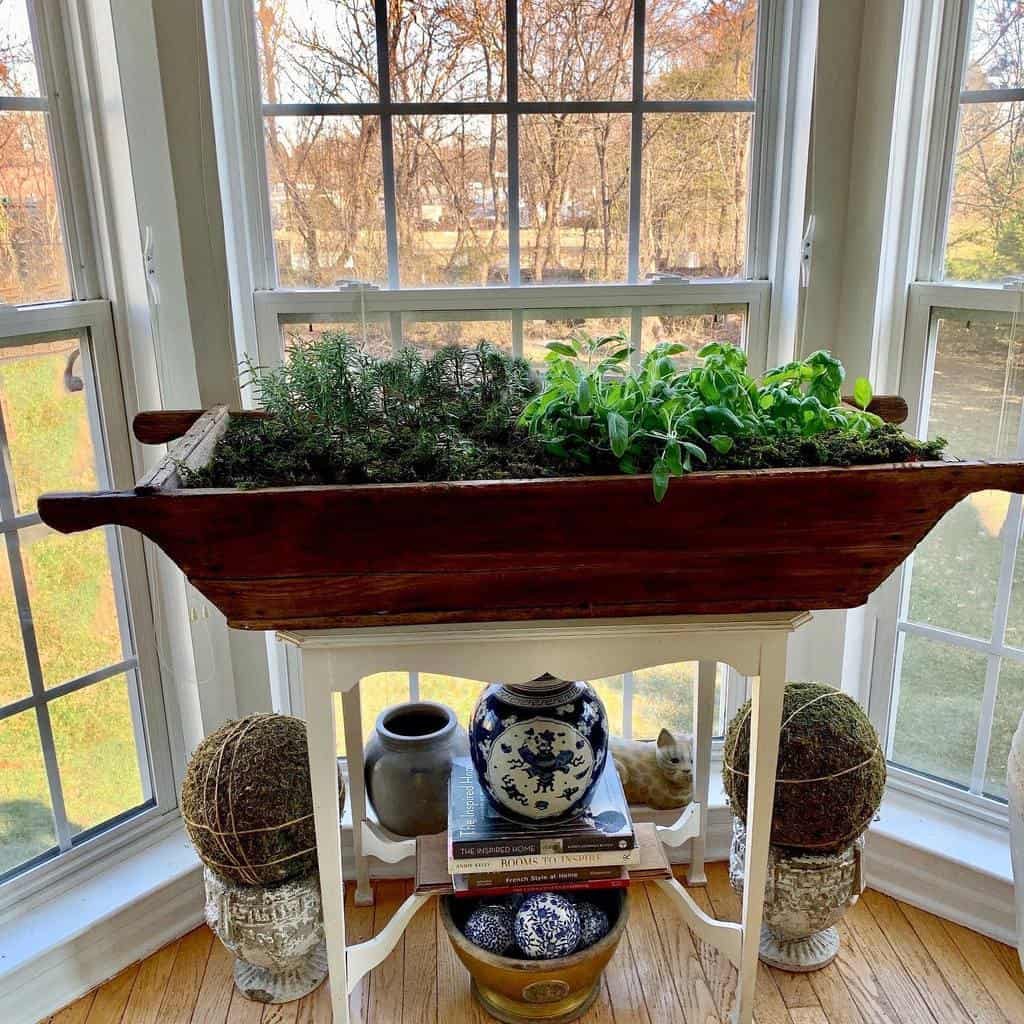 A wooden planter with herbs on a white stand by a window, surrounded by decorative items and books