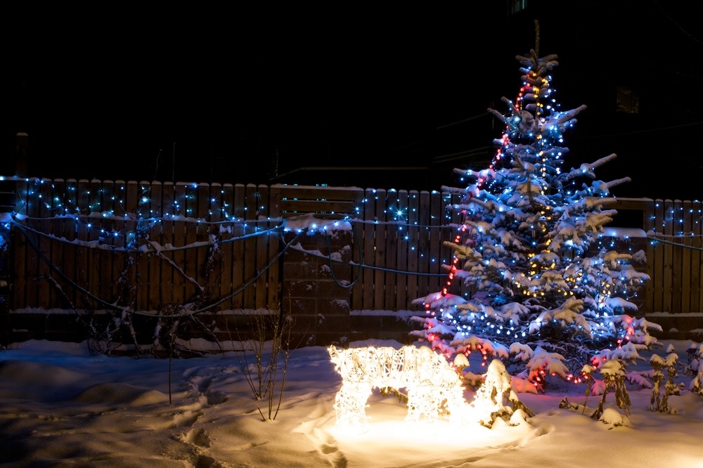 Snow-covered yard at night with illuminated Christmas tree and a lit deer figure, string lights adorn a wooden fence