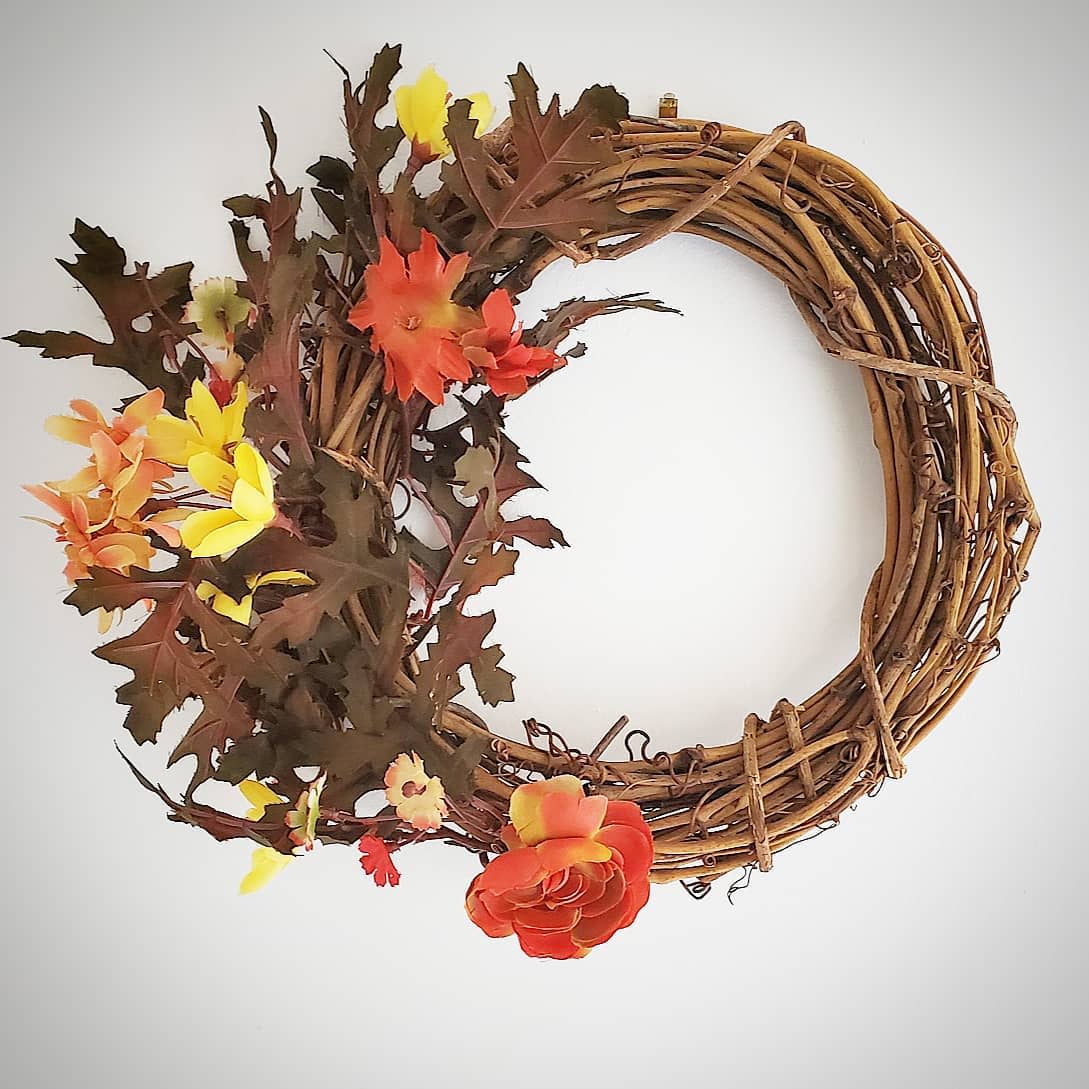 A decorative fall wreath made of intertwined branches, adorned with orange and yellow flowers and green leaves against a white background