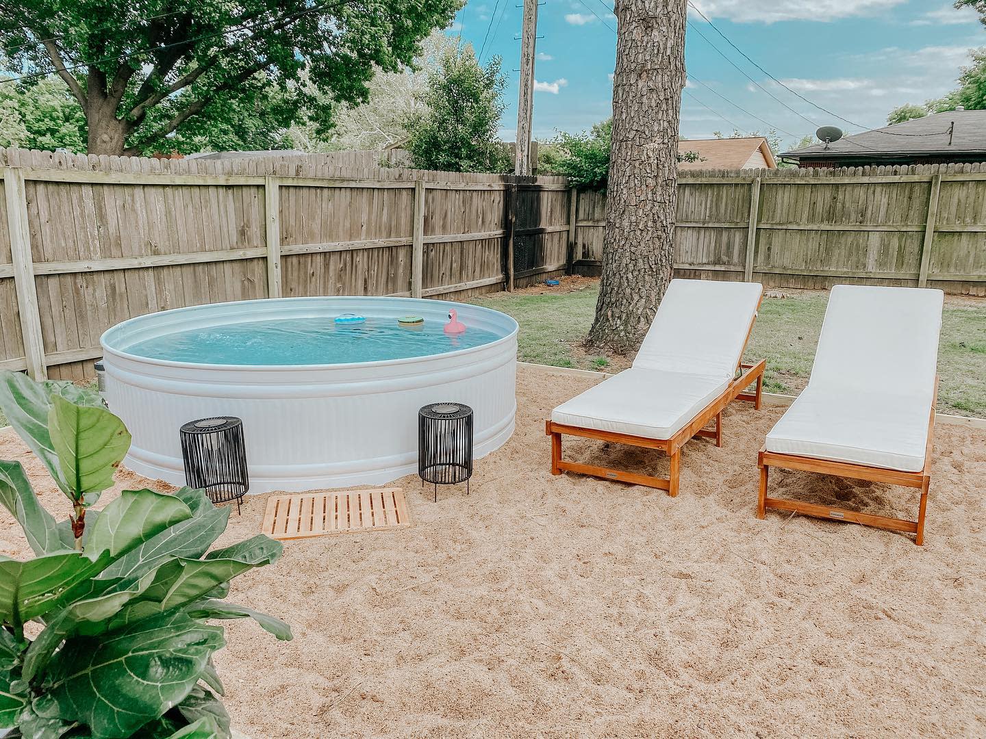 Serene backyard with a white stock tank pool, sandy lounge area, wooden sun loungers, black lanterns, and a tall wooden fence