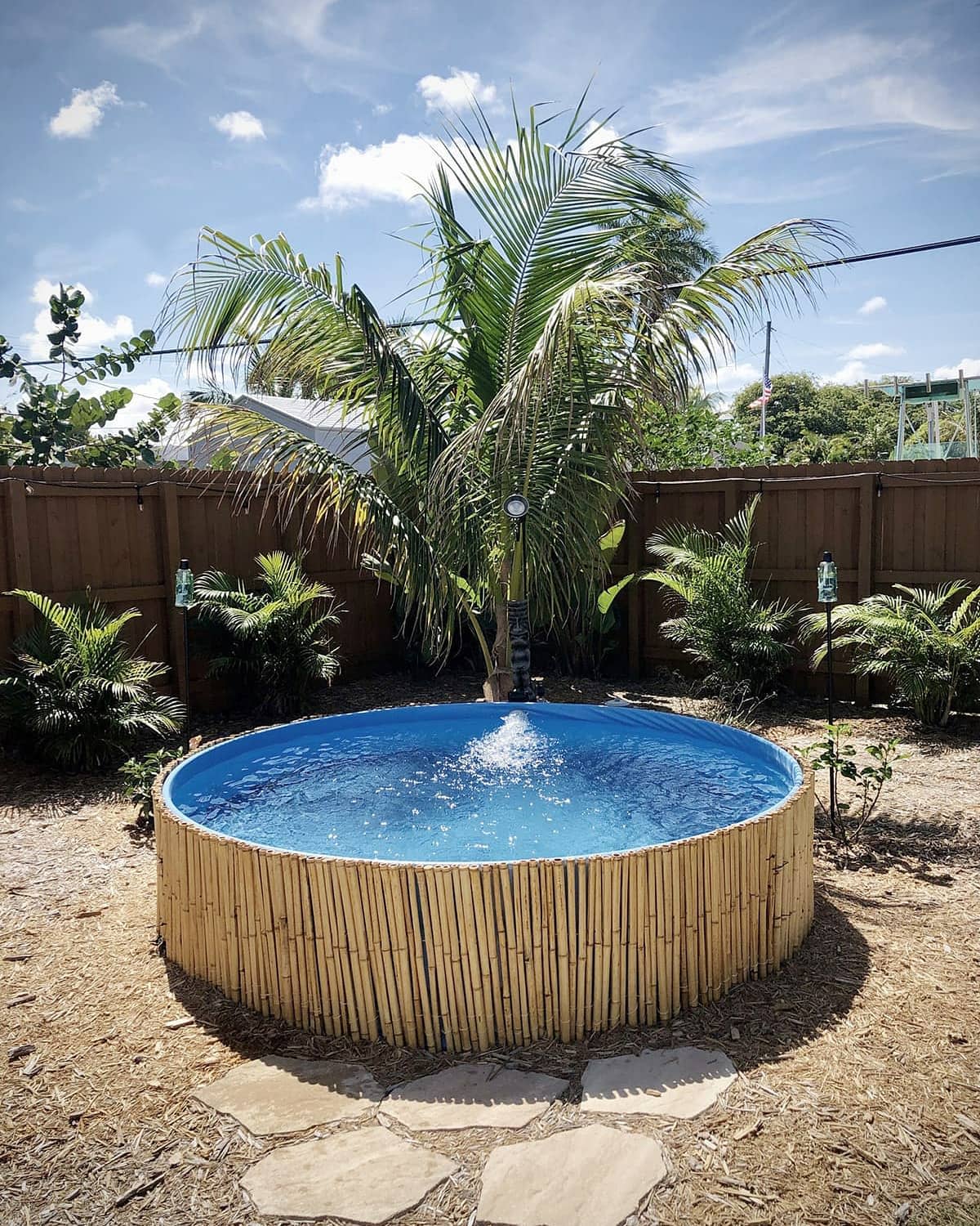 Tropical backyard with a stock tank pool wrapped in bamboo, surrounded by palm trees, lush greenery, and a wooden fence under a blue sky