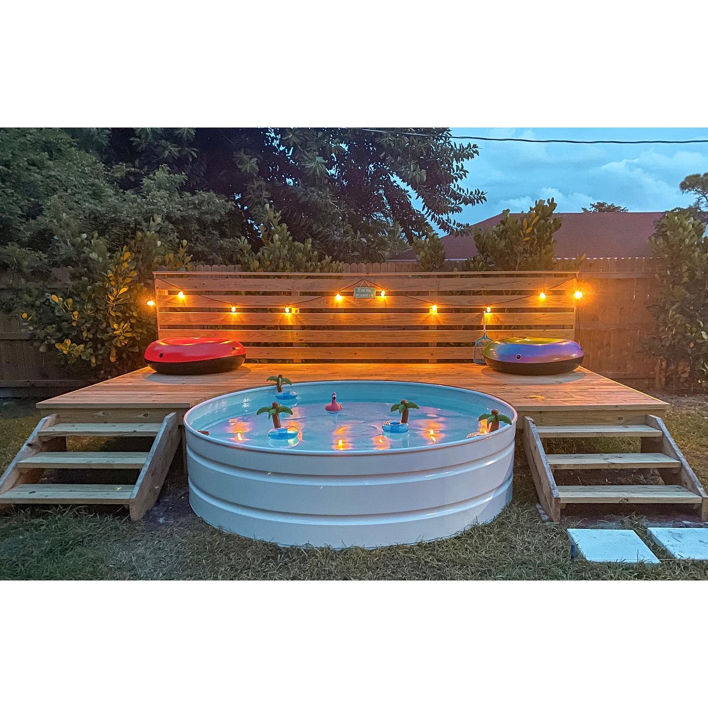 A round above-ground pool with inflatables on a wooden deck, surrounded by string lights, with steps leading to the deck and trees in the background