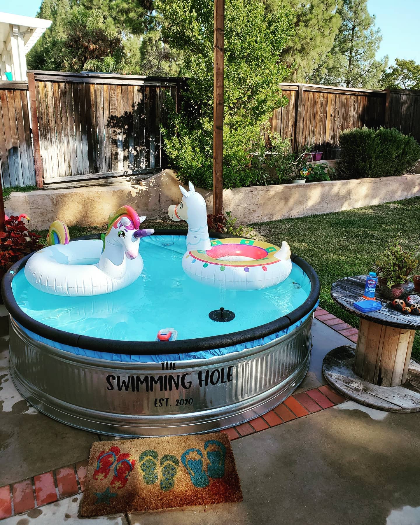 A backyard setup with a round pool featuring unicorn and llama floats, a small table with drinks, and a fenced grassy area