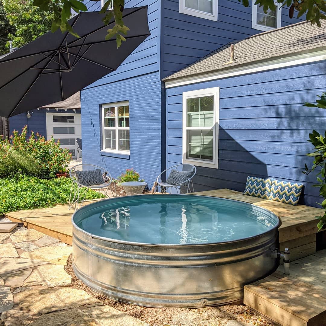 Outdoor stock tank pool surrounded by a wooden deck, chairs, and a large umbrella, next to a blue house with white-trimmed windows