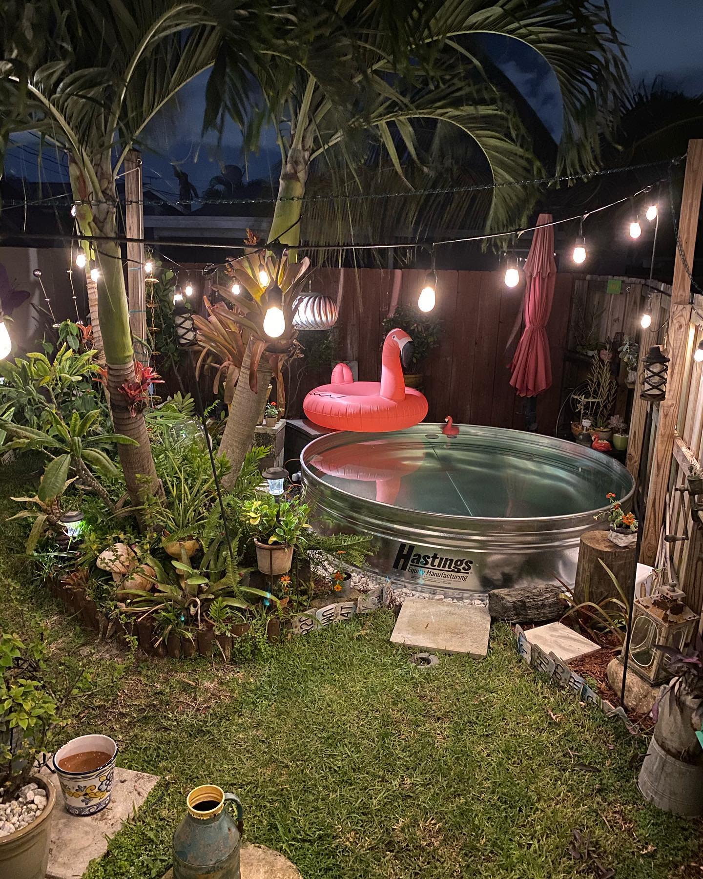 Cozy backyard scene with a round metal pool, pink flamingo float, string lights, lush plants, and garden decor at night