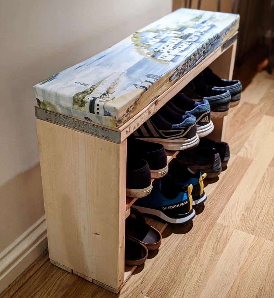 Wooden shoe rack with three shelves, holding various sneakers and flip-flops, topped with a cushion in a hallway