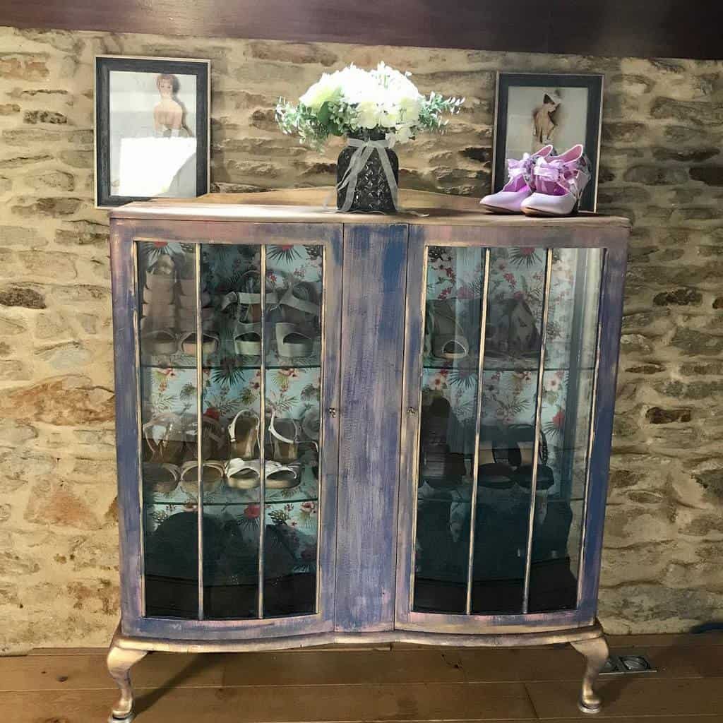 A vintage cabinet with glass doors displaying shoes, topped with a vase of flowers and framed photos, set against a stone wall