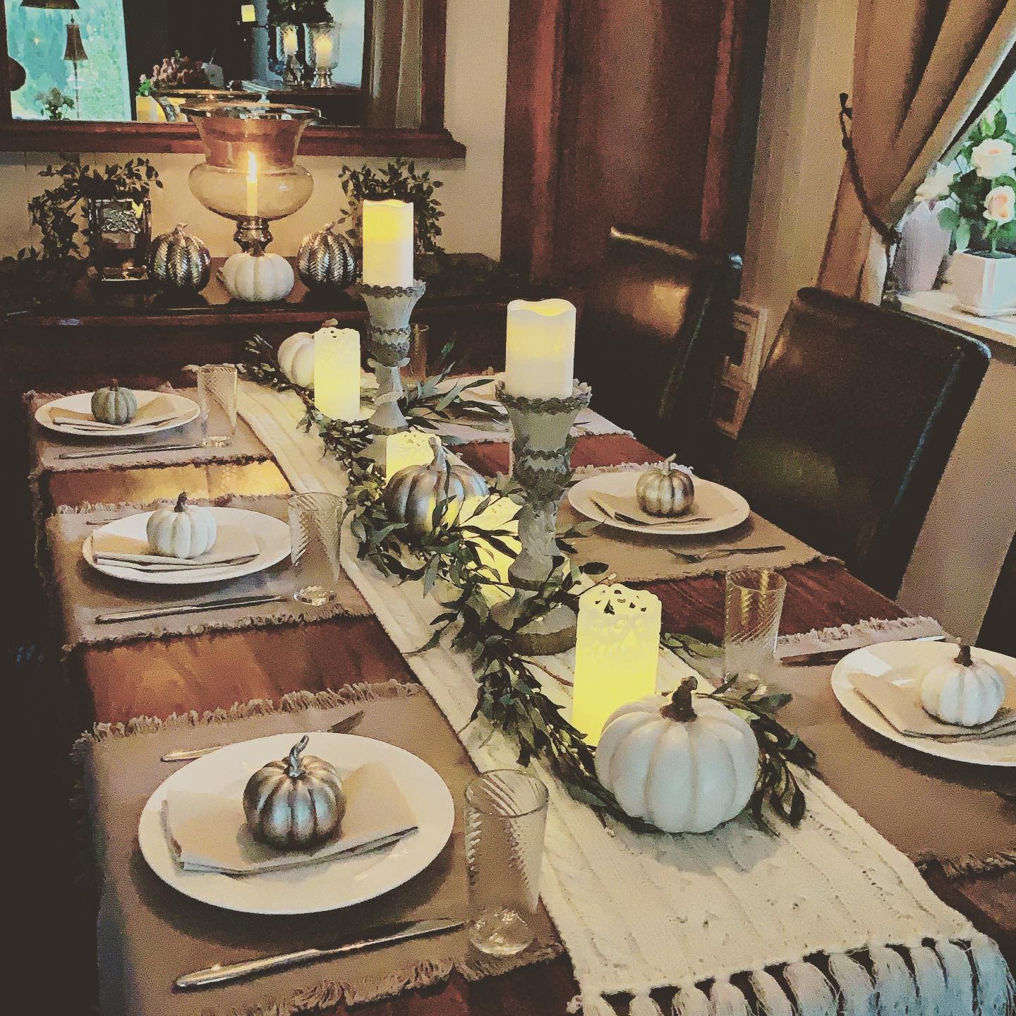 Cozy dining table with white pumpkins, candles, and greenery centerpiece; plates and cutlery are set for a meal creating a warm, inviting ambiance