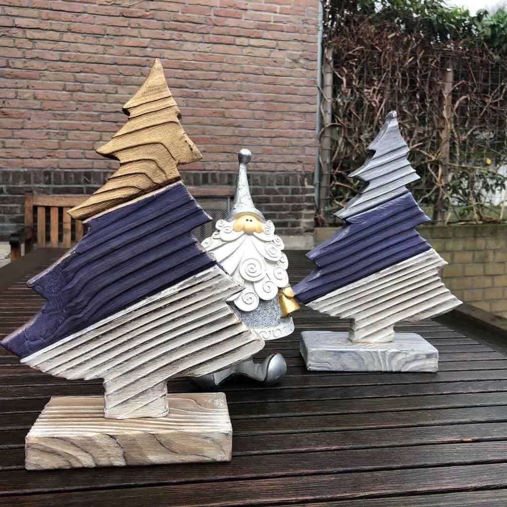 Wooden Christmas tree decorations and a gnome figurine on a wooden table outside, with a brick wall and bushes in the background