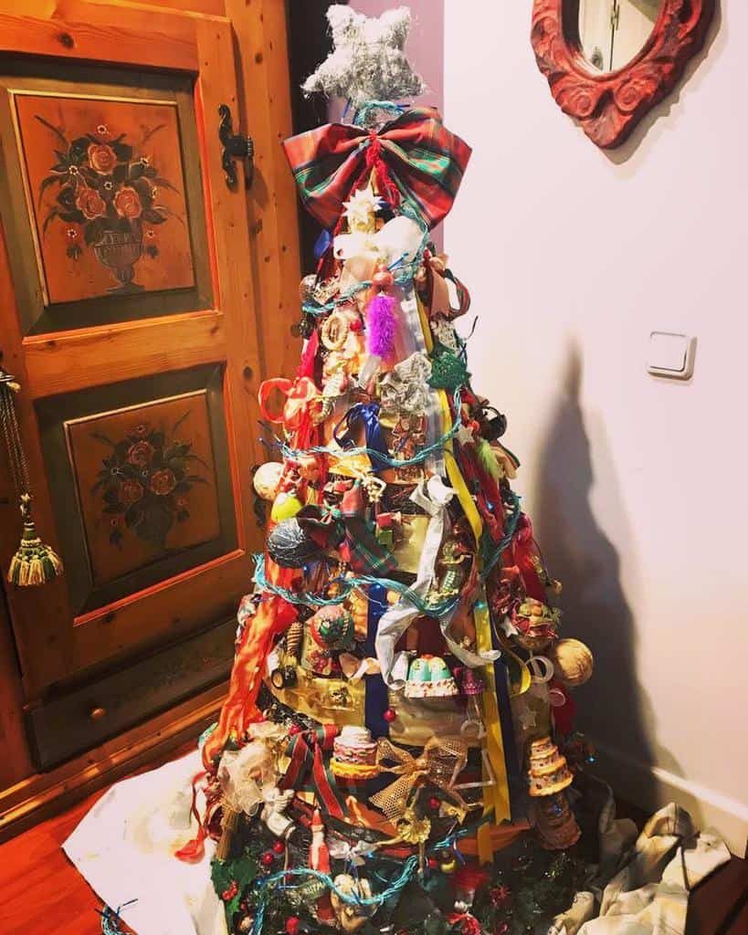 A colorful Christmas tree decorated with various ornaments, ribbons, and a star on top, standing beside a wooden door