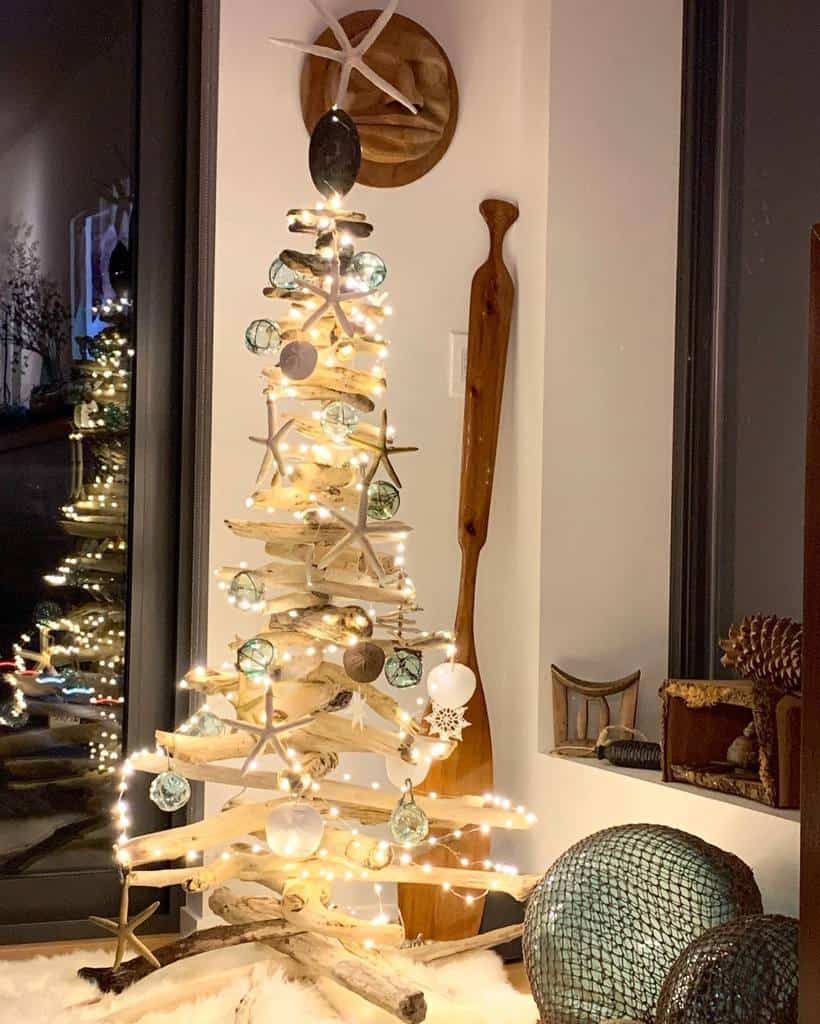 A driftwood Christmas tree decorated with lights and ornaments stands near a wooden paddle and a window