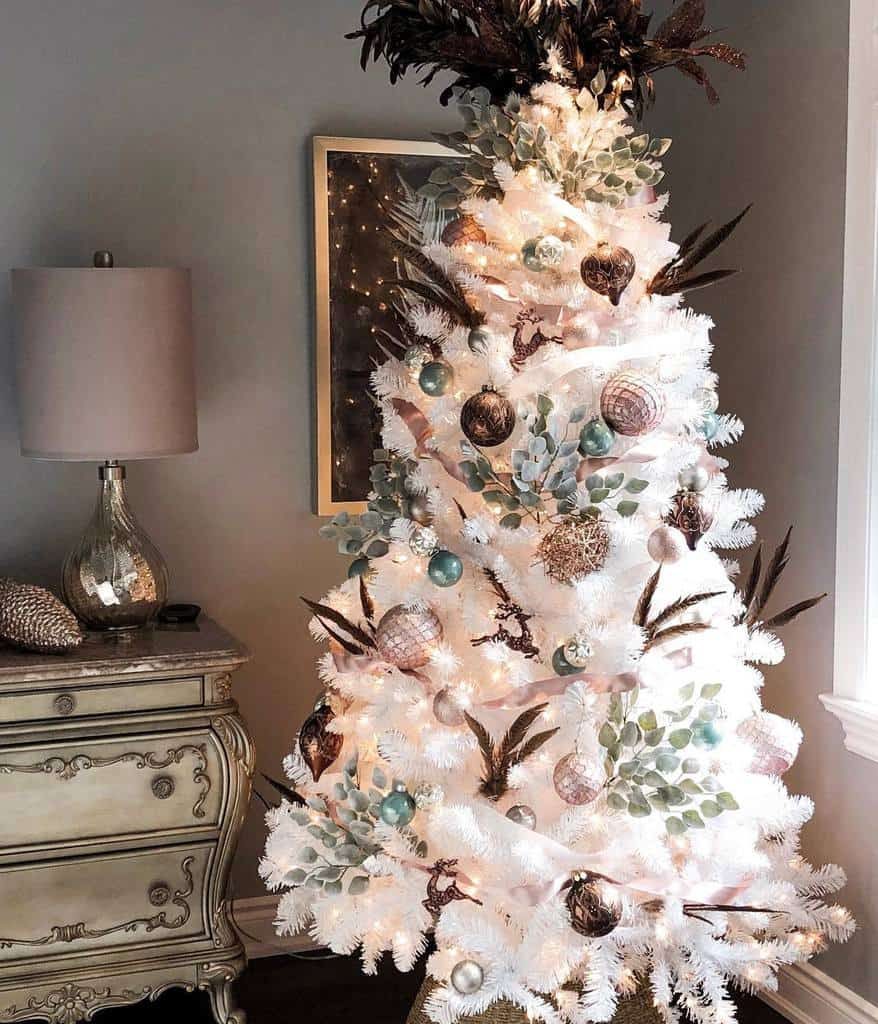 White Christmas tree with silver and teal ornaments, brown feathers, and lights, placed near a decorative lamp and ornate dresser