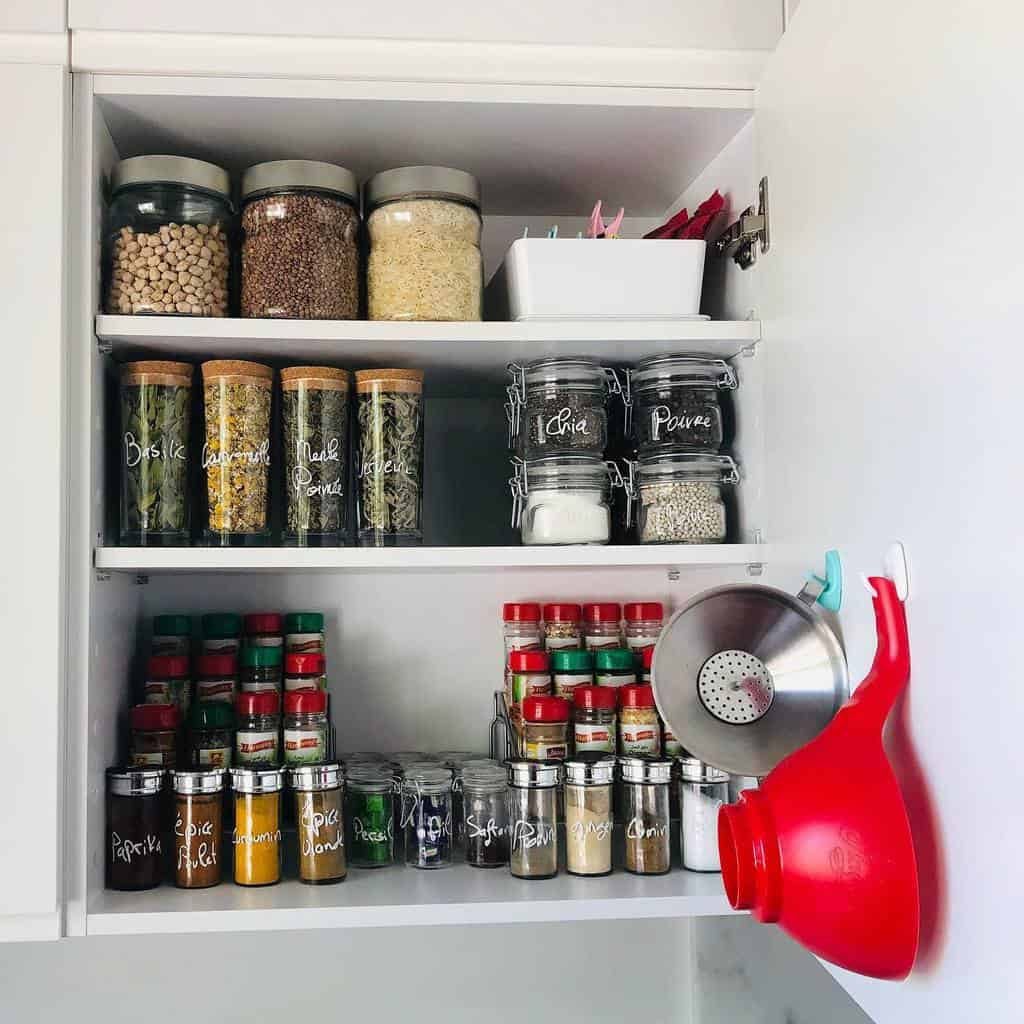 An organized spice cabinet with labeled jars and bottles filled with various spices and grains, with a funnel hanging on the right side