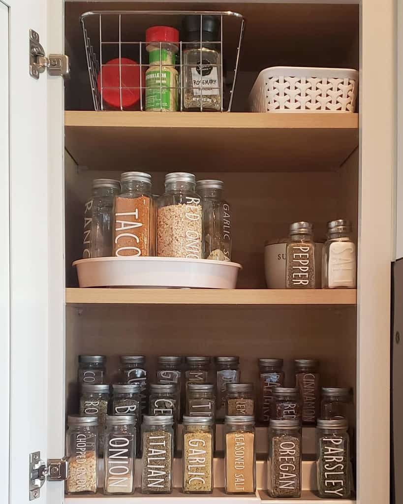 Organized spice cabinet with labeled jars containing various spices, bottles of oil, and a basket with containers on a wooden shelf