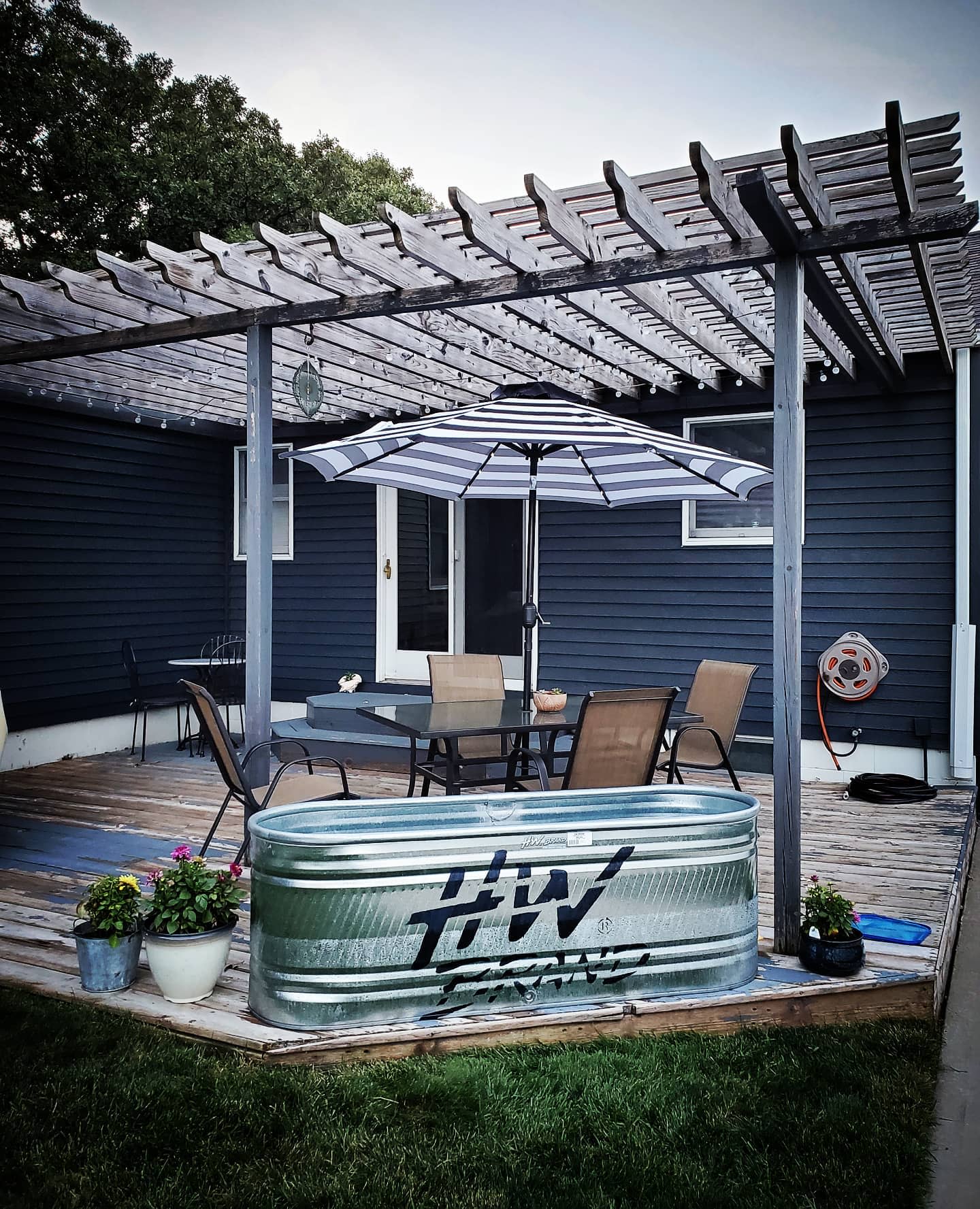 Galvanized stock tank pool on a wooden deck with a pergola, outdoor dining set, striped umbrella, and potted flowers in a cozy backyard