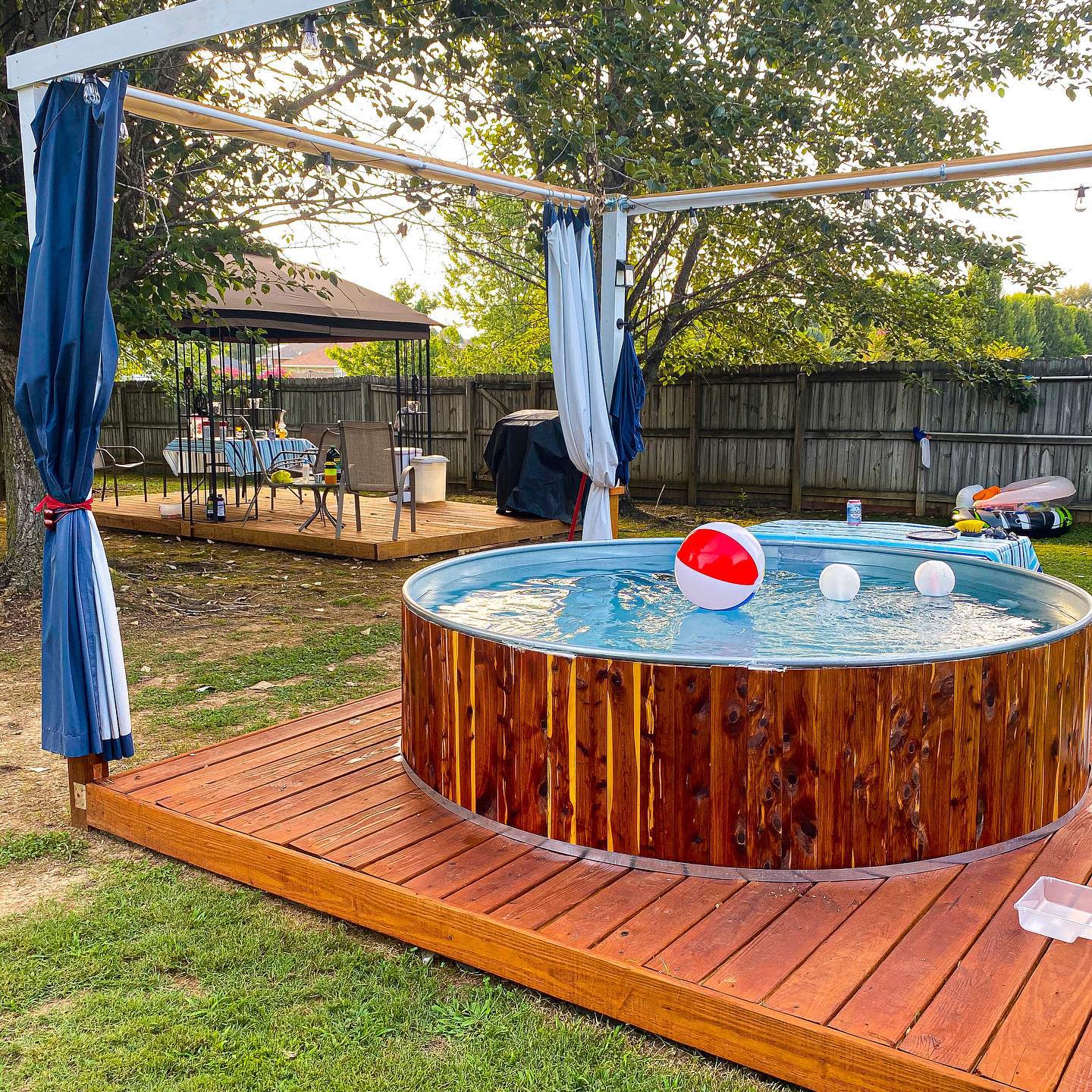 Backyard stock tank pool with a wood-paneled exterior, raised deck, pergola with curtains, and a nearby outdoor dining area