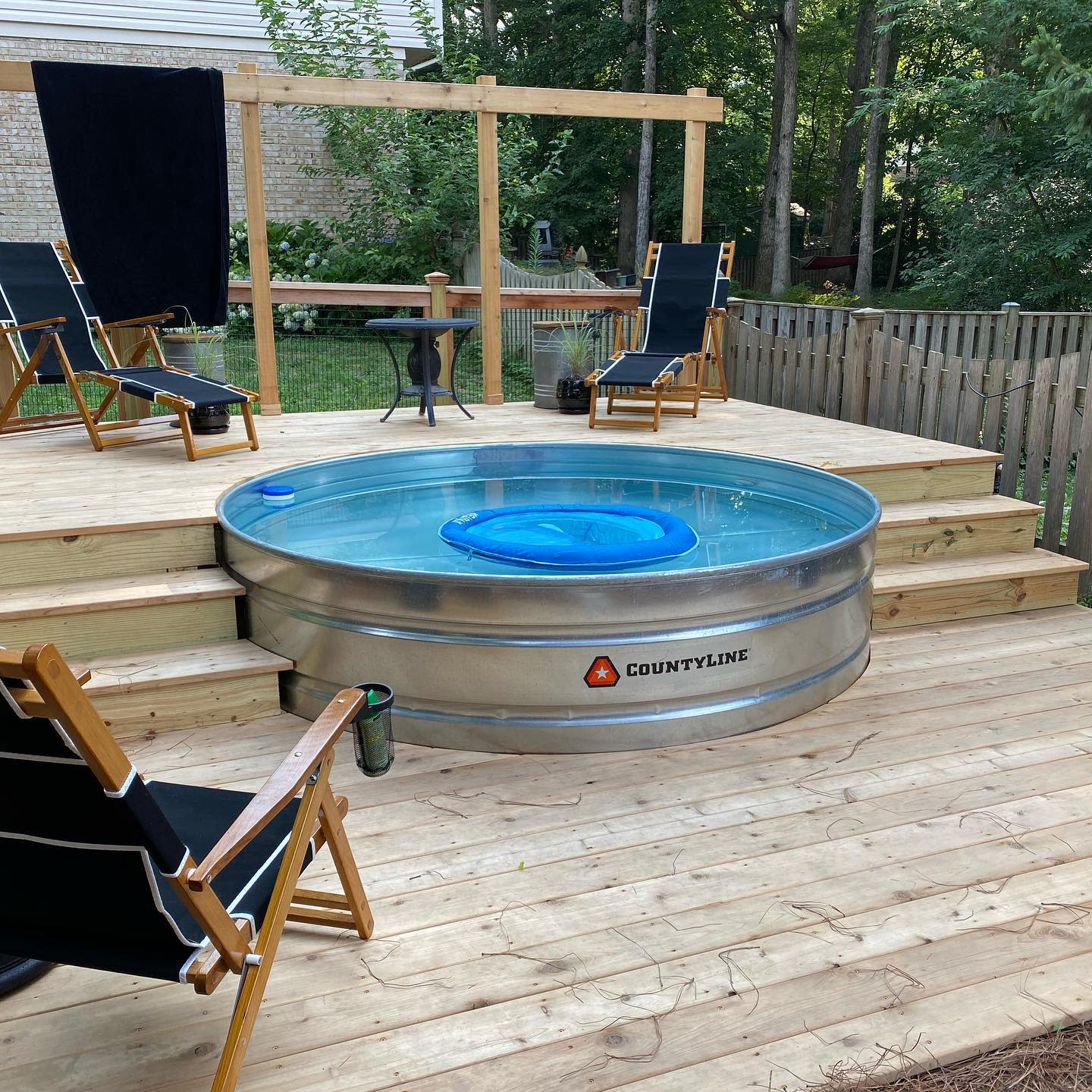 A round pool on a wooden deck with black chairs and a small table around it, set in a backyard with trees