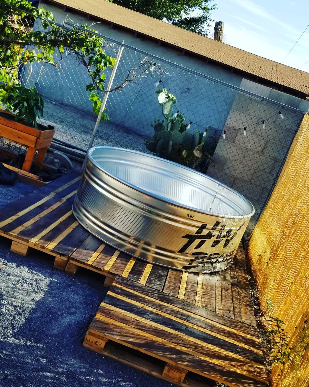 Galvanized stock tank pool on a rustic wooden pallet deck in a backyard with a chain-link fence, cacti, and string lights