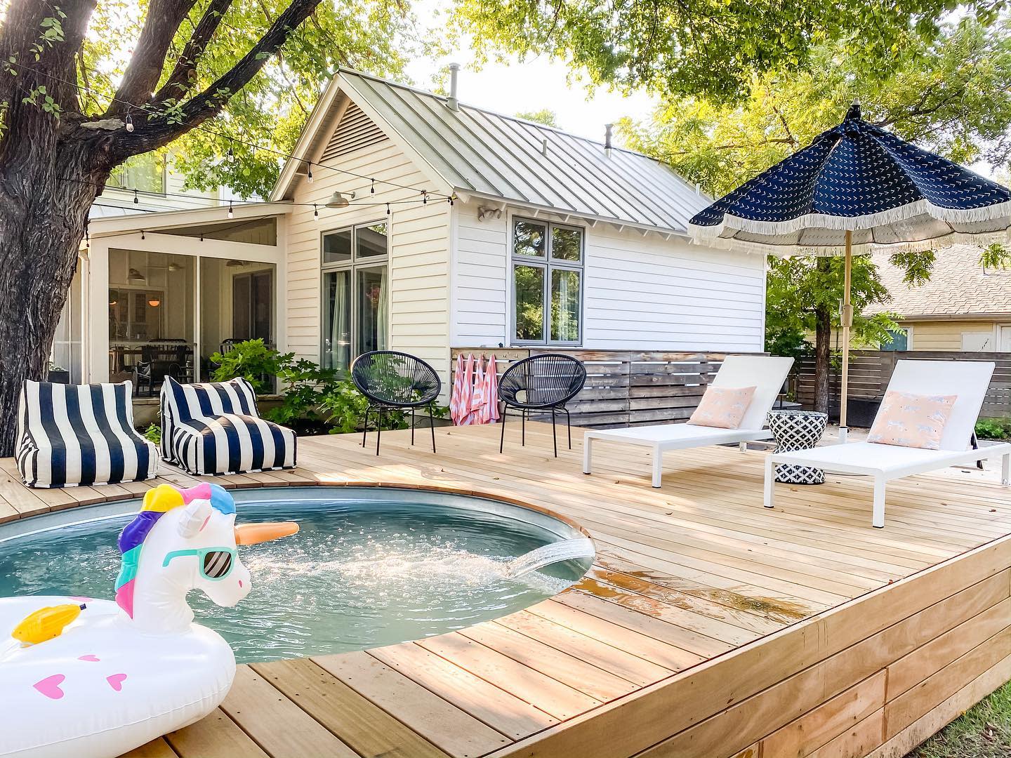 A small above-ground pool with a unicorn float in a backyard with striped lounge chairs, umbrella, and a white house in the background