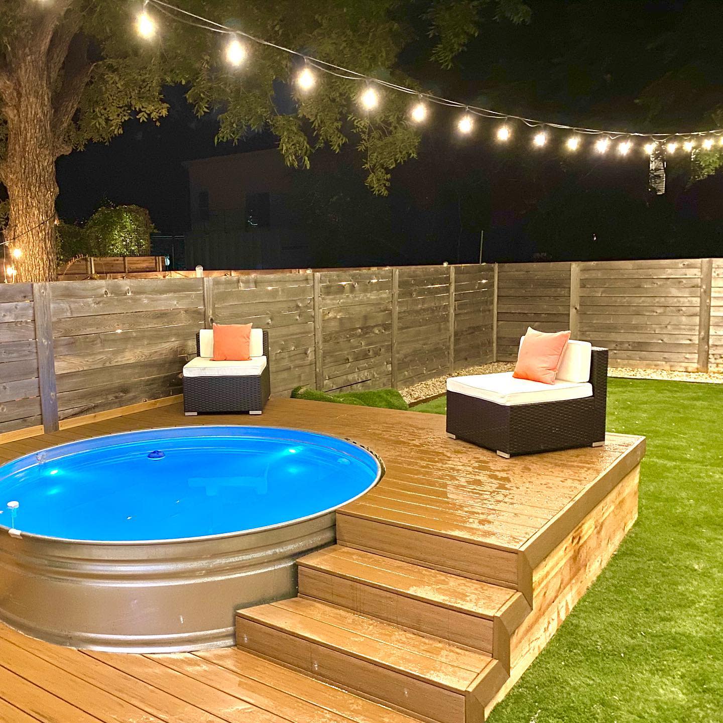 Modern backyard with a stock tank pool on a wooden deck, cozy lounge seating, string lights, and a dark wood privacy fence at night