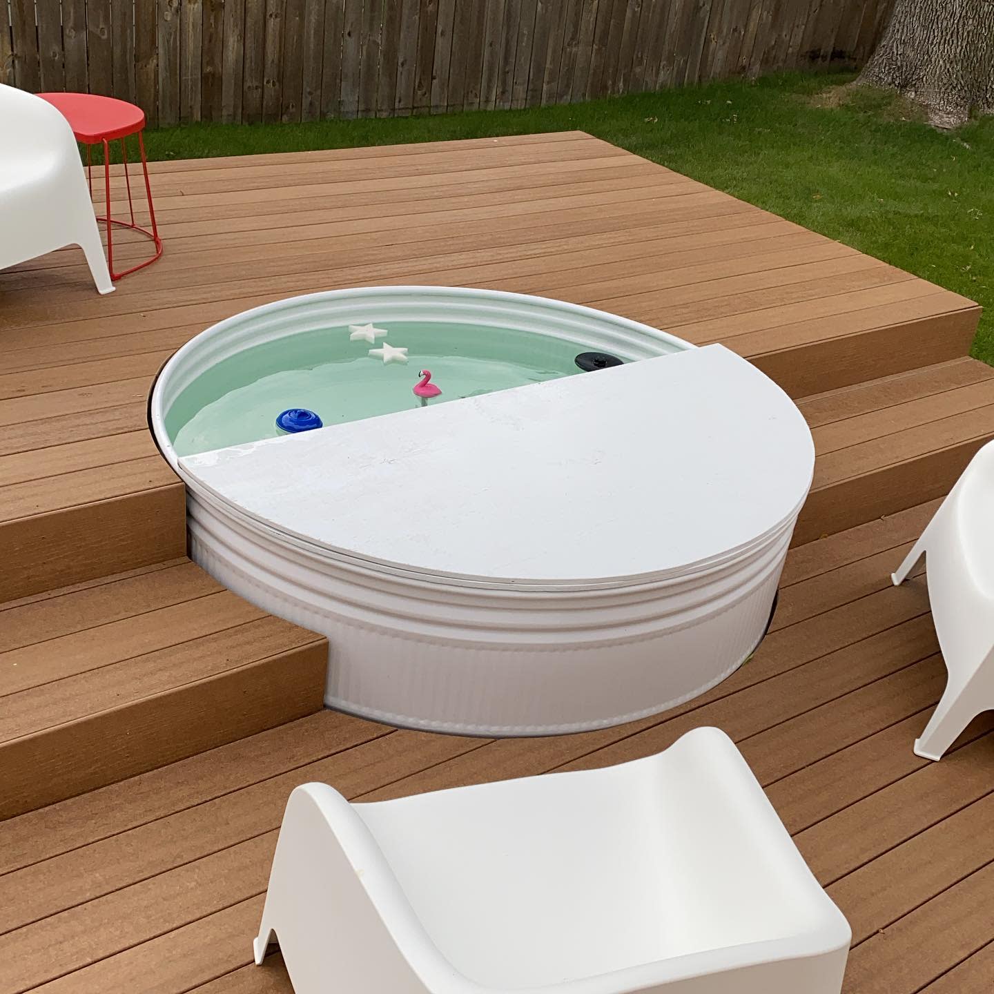 Modern backyard deck with a white stock tank pool partially covered, sleek white chairs, and floating toys for a stylish outdoor retreat