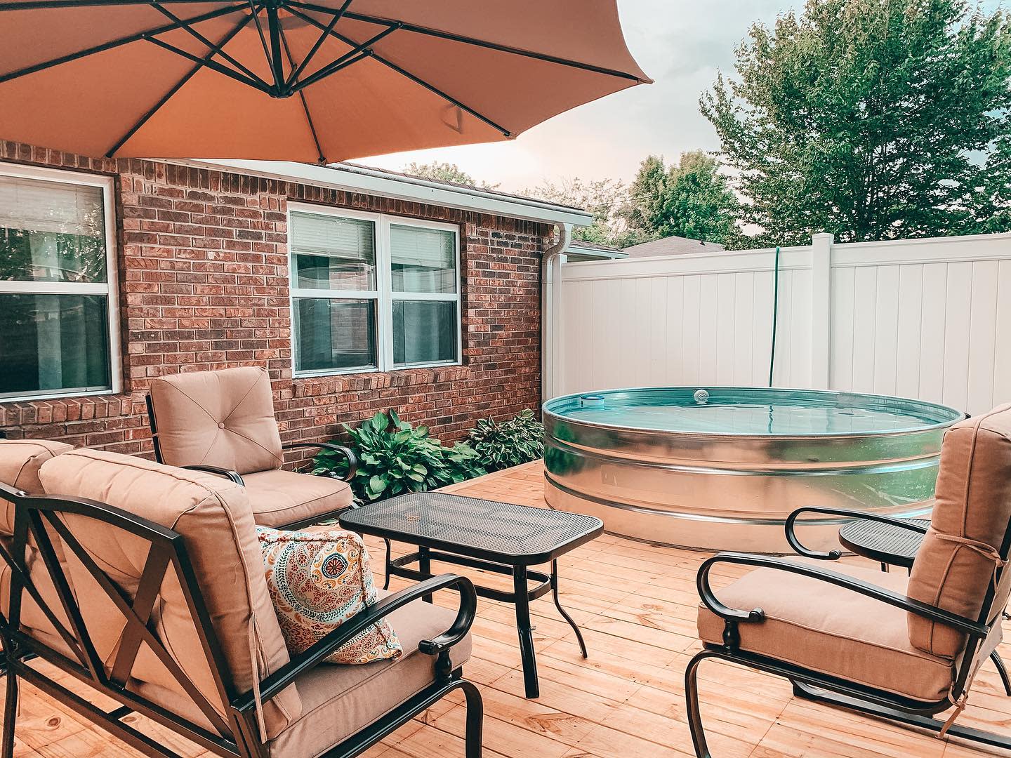 Patio with cushioned chairs, table, large umbrella, and a round stock tank pool on a wooden deck beside a brick house and white fence