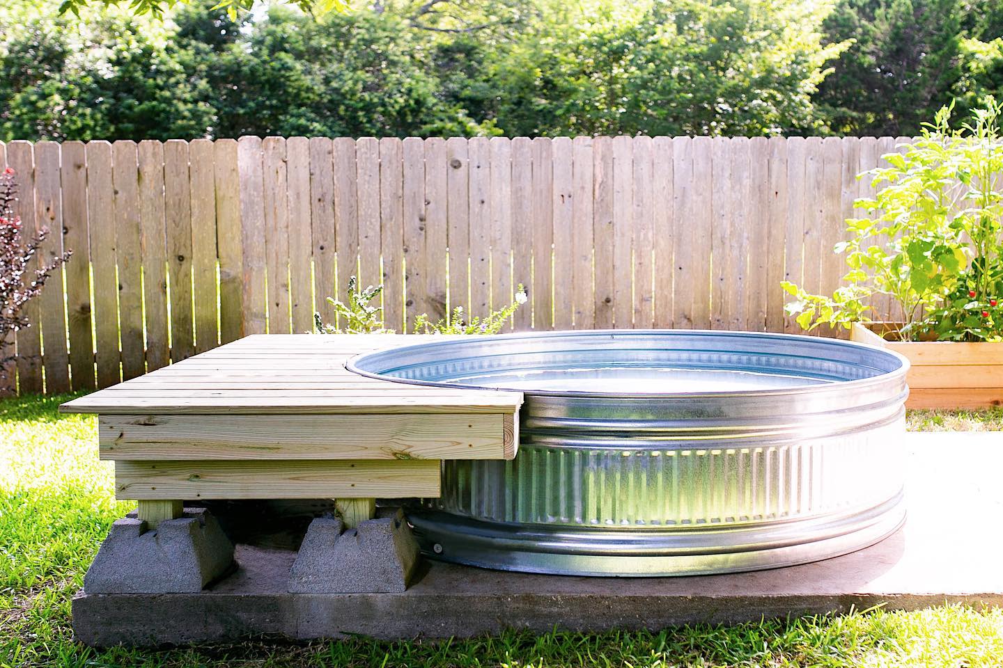 Metal stock tank pool with wooden deck in a sunny backyard, surrounded by a wooden fence and greenery