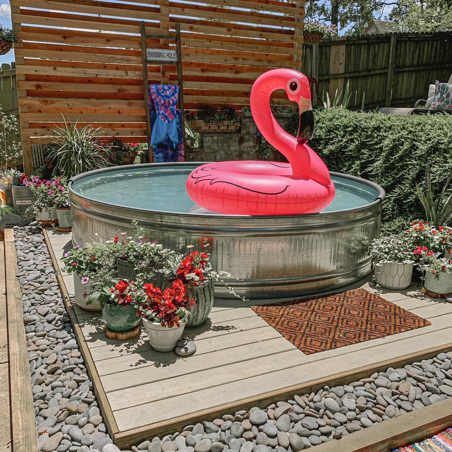 A backyard stock tank pool with a pink flamingo float, surrounded by potted plants and a wooden privacy screen