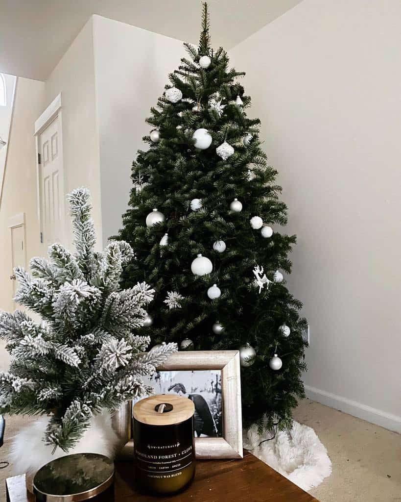 A decorated Christmas tree with white ornaments, a small frosted tree, a candle, frames, and a faux fur rug on the floor