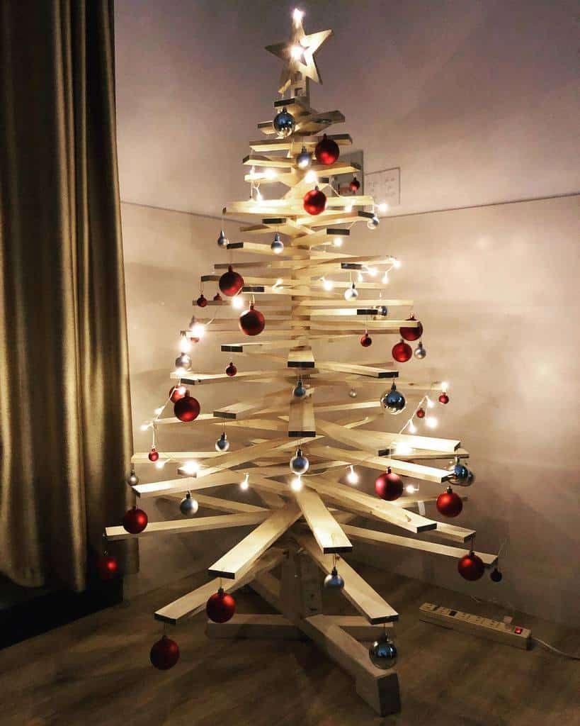 Wooden Christmas tree with red and blue ornaments and warm white lights, set in a dimly lit room with a curtain on the left