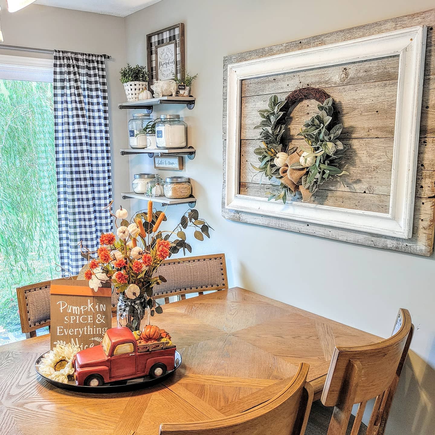 Cozy dining area with fall decor: wooden table, rustic shelves, pumpkin spice sign, red truck centerpiece, and autumn wreath on wall