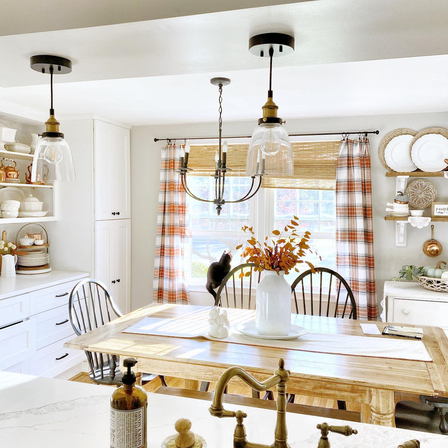 Bright kitchen with white cabinets, a wooden table set with chairs, plaid curtains, pendant lights, and a vase of autumn leaves