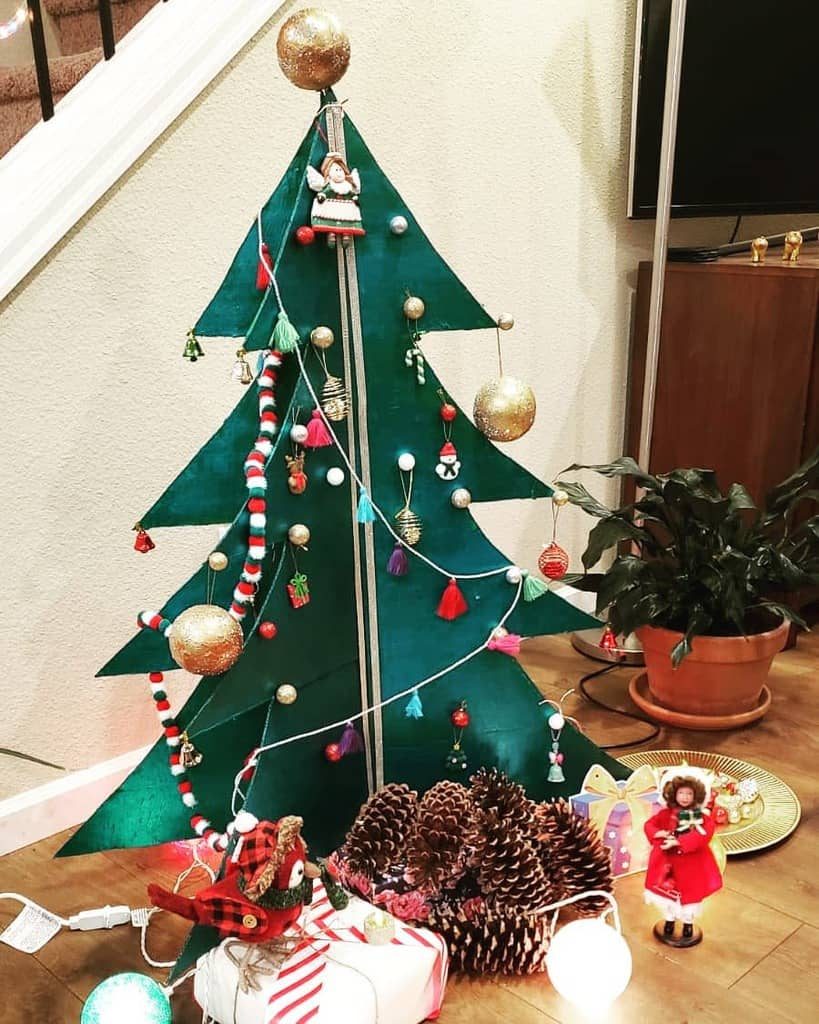 A triangular Christmas tree with ornaments, large pinecones, presents, and a doll sits on the floor near a potted plant