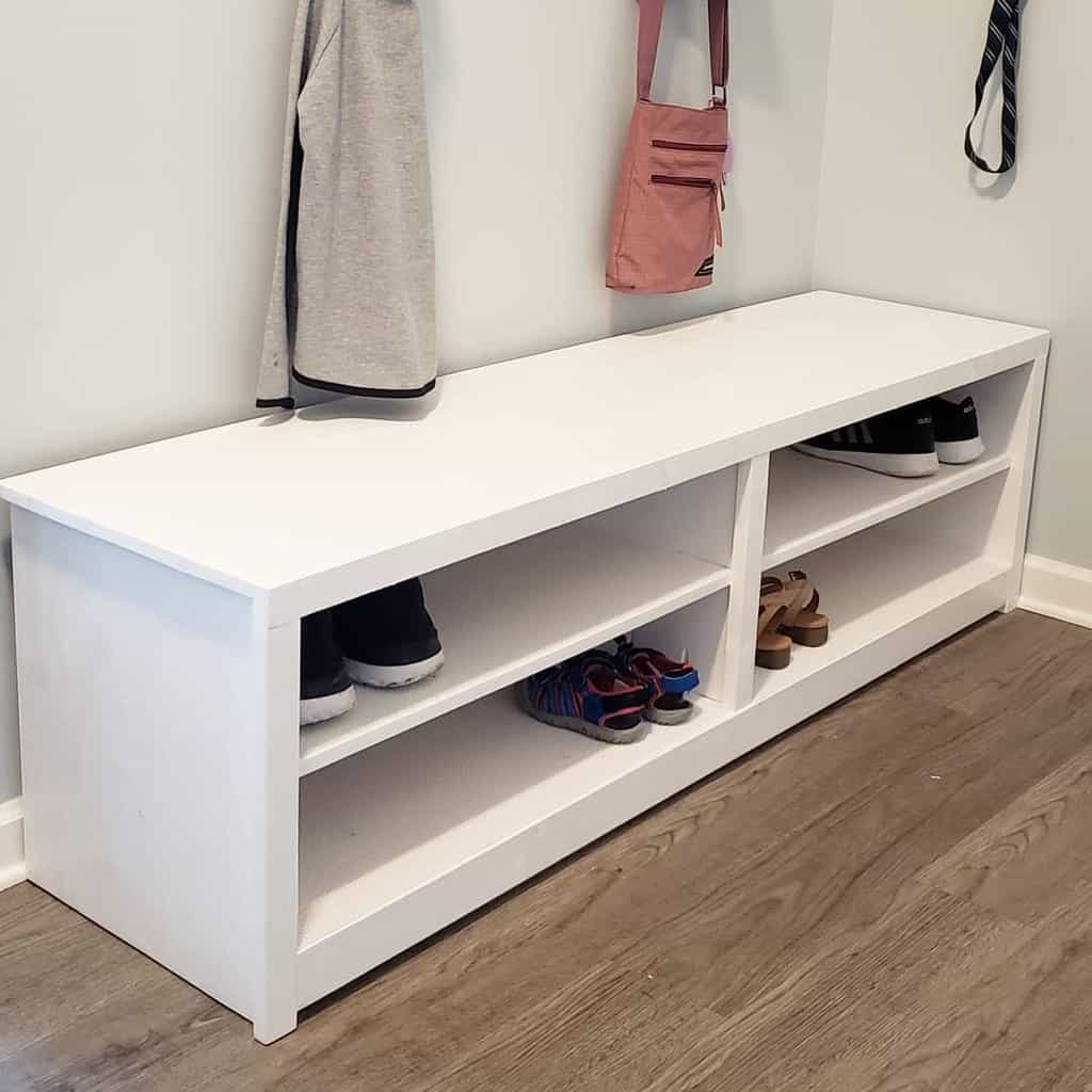 White wooden shoe storage bench with open shelves, neatly organizing footwear in a mudroom with hanging coats and bags