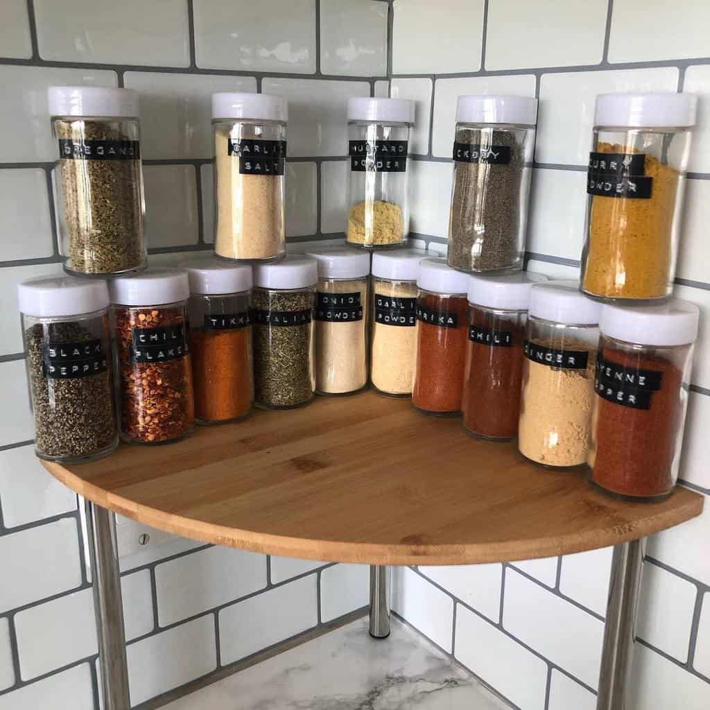 A collection of labeled spice jars arranged on a rounded wooden shelf in a tiled kitchen corner