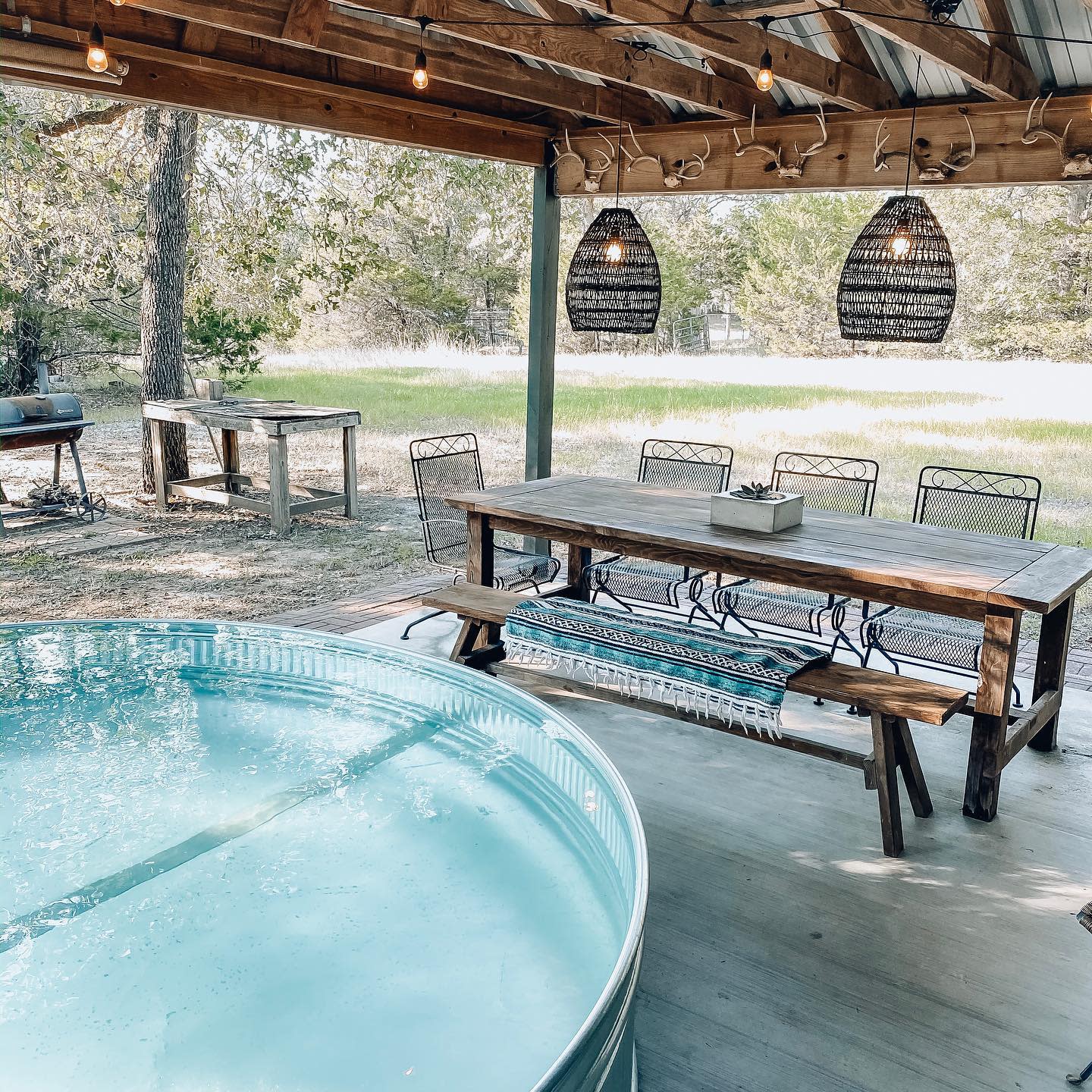 Outdoor patio with a wooden dining table, benches, and a metal pool; string lights hang above, with trees and grass in the background