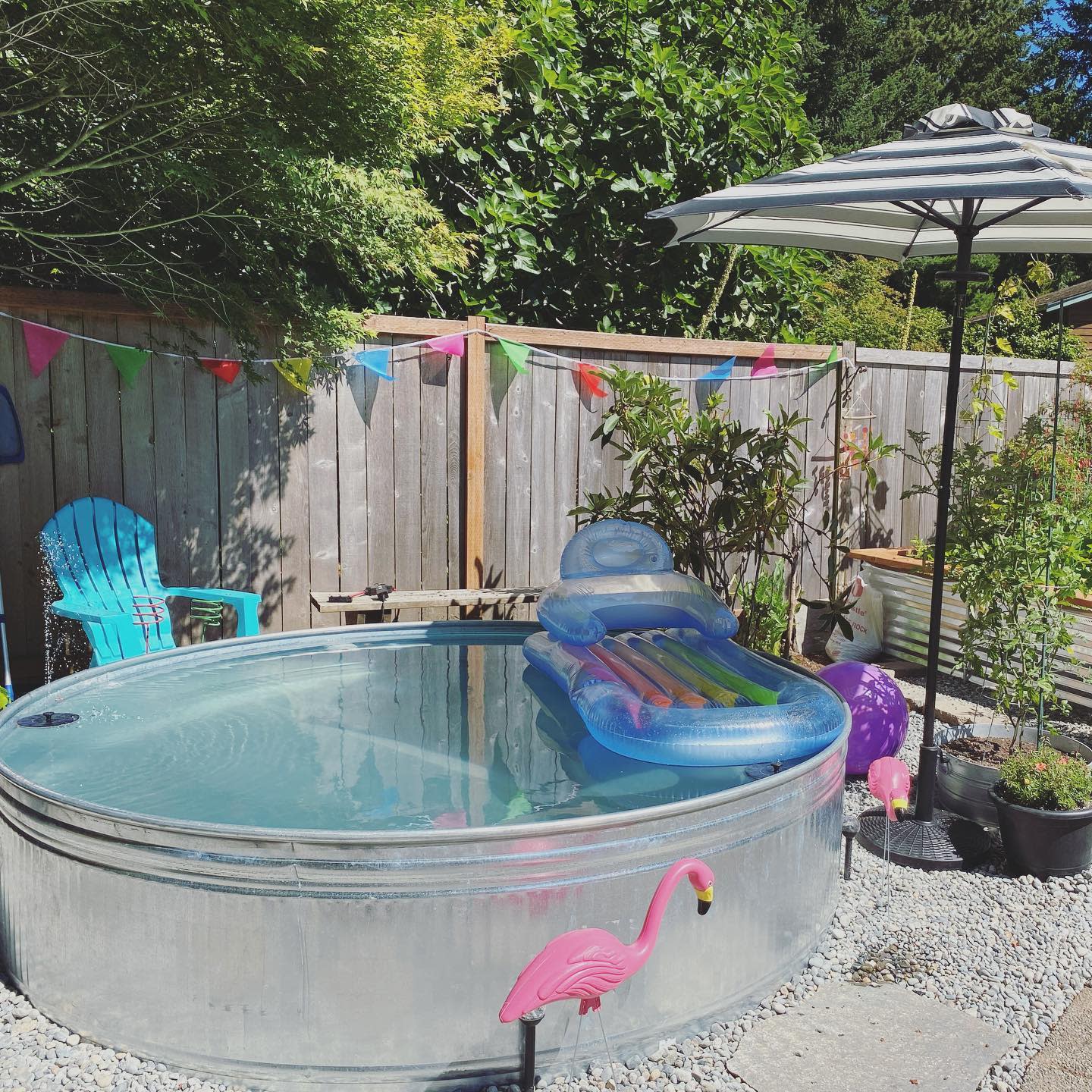 Circular pool with an inflatable lounge, surrounded by flamingo decorations, a blue chair, umbrella, and colorful bunting in a garden