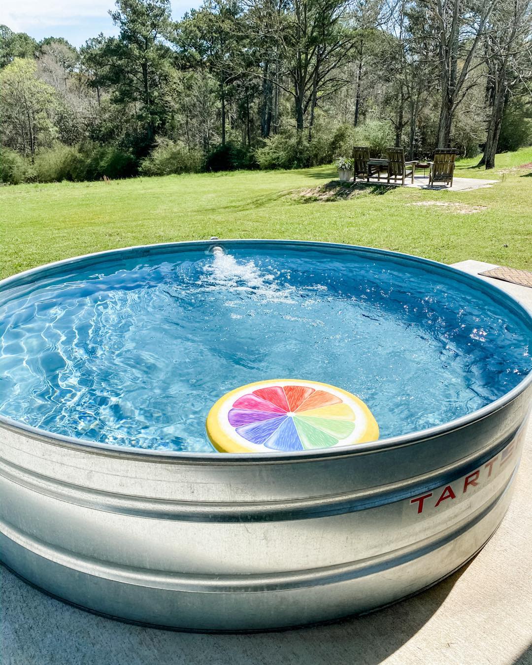 A small round pool with a colorful inflatable, set in a grassy yard with trees and a wooden seating area in the background