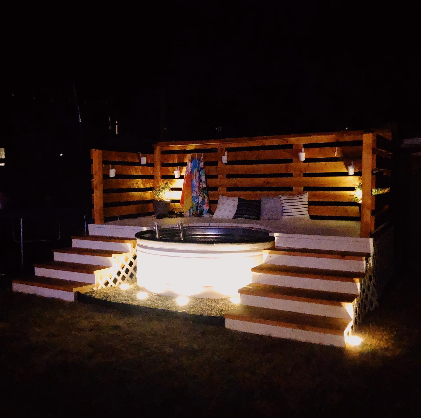 Wooden deck with a hot tub, colorful towels, and striped cushions, softly illuminated by outdoor lights at night