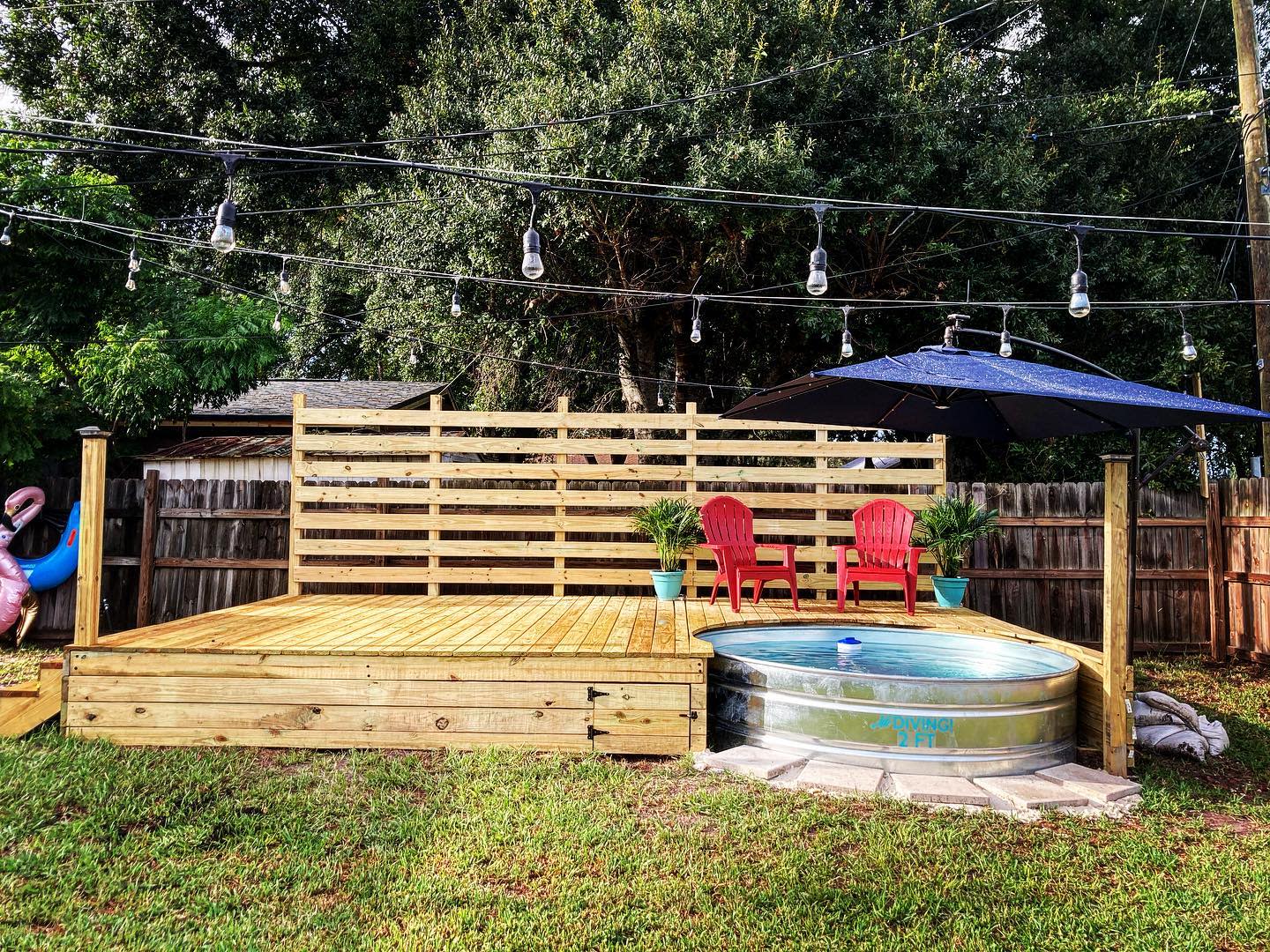 Wooden deck with string lights, two red chairs, a small pool with umbrella, and greenery in the background