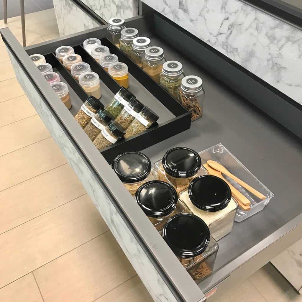 Open kitchen drawer with neatly organized spice jars, containers, and wooden utensils on a marble background