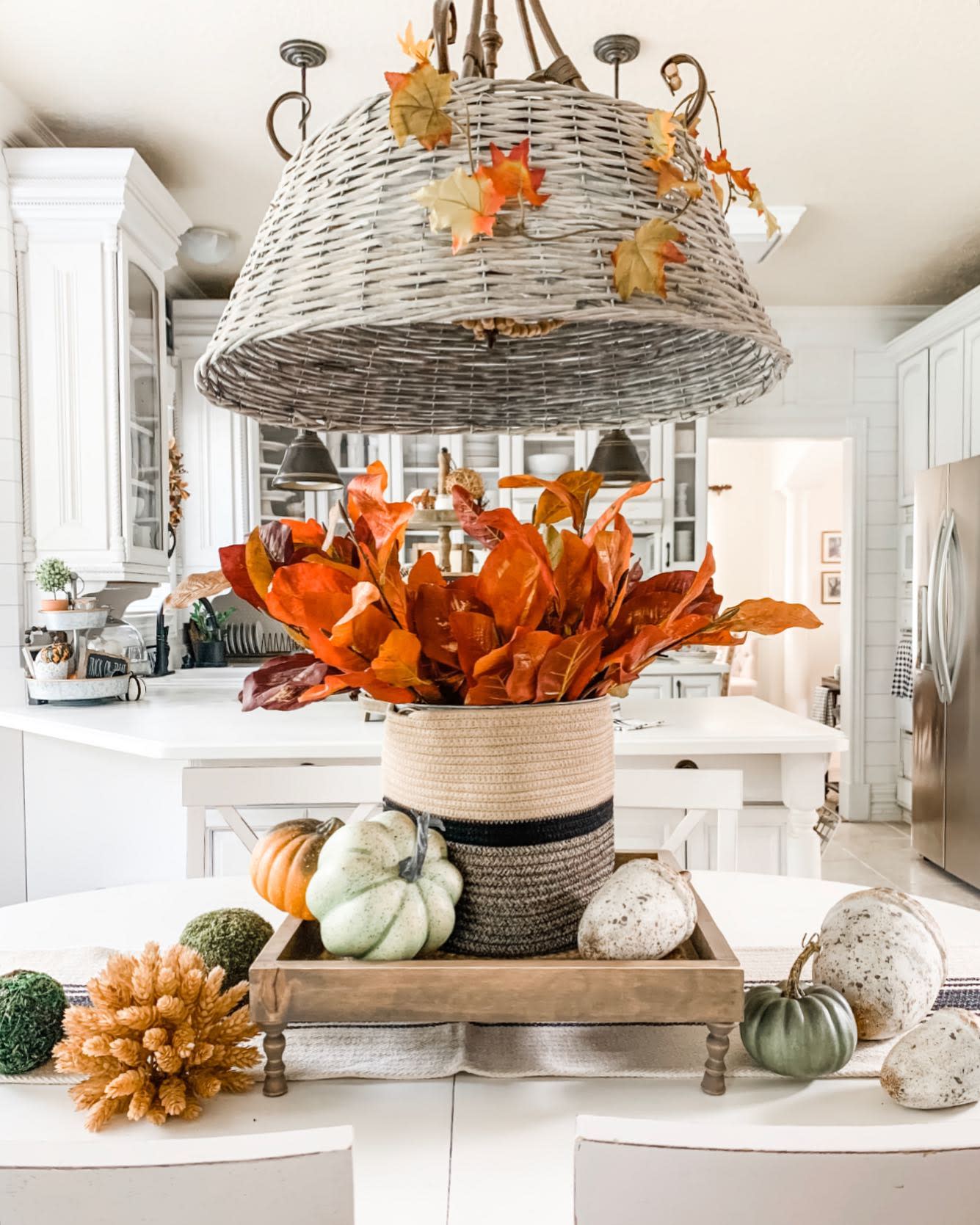 Kitchen with a fall-themed table display featuring pumpkins, a basket with orange leaves, and a wicker chandelier with autumn leaves