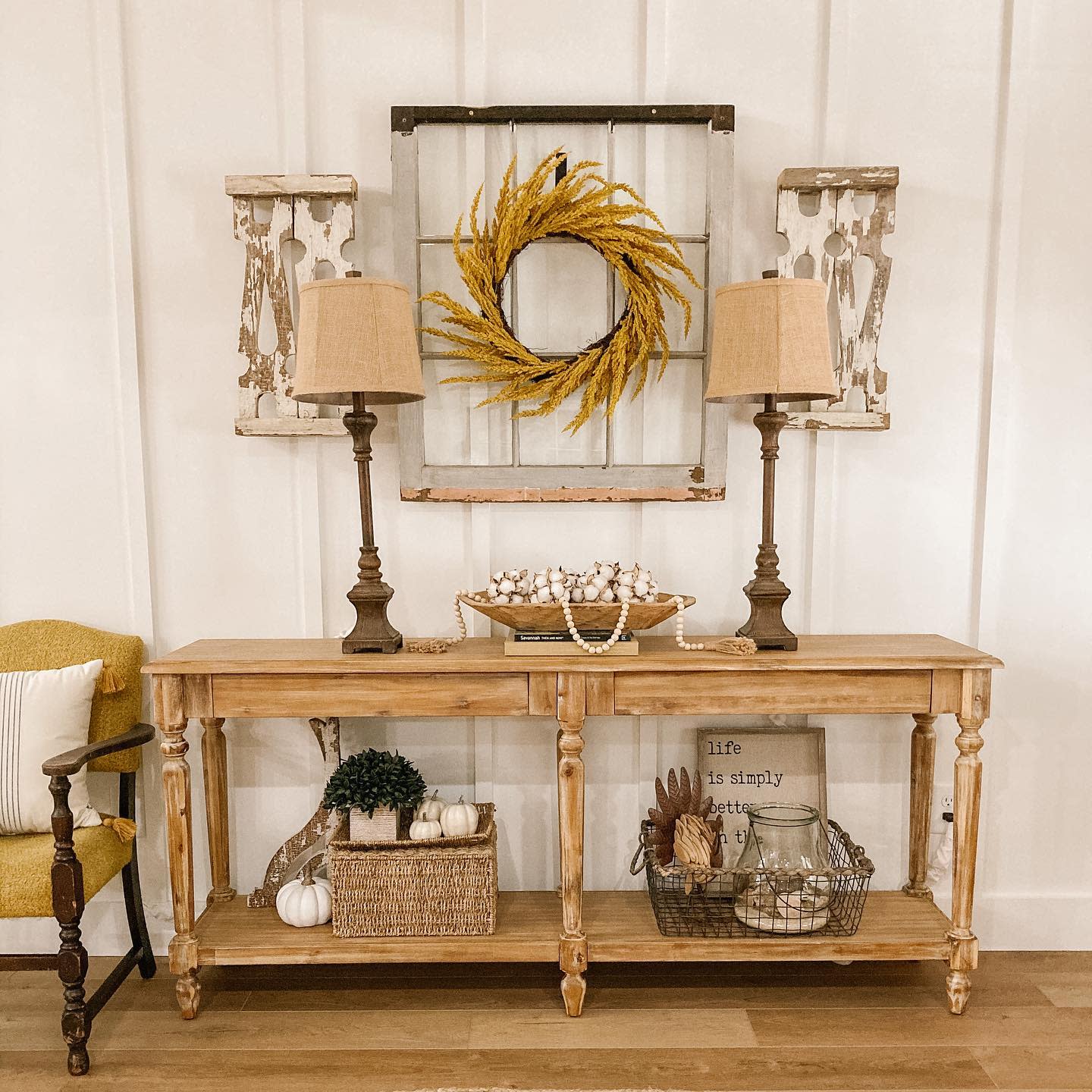 Rustic wooden console table with two lamps, a wreath, and decor items like baskets and pumpkins on a wood-paneled wall, creating a cozy ambiance