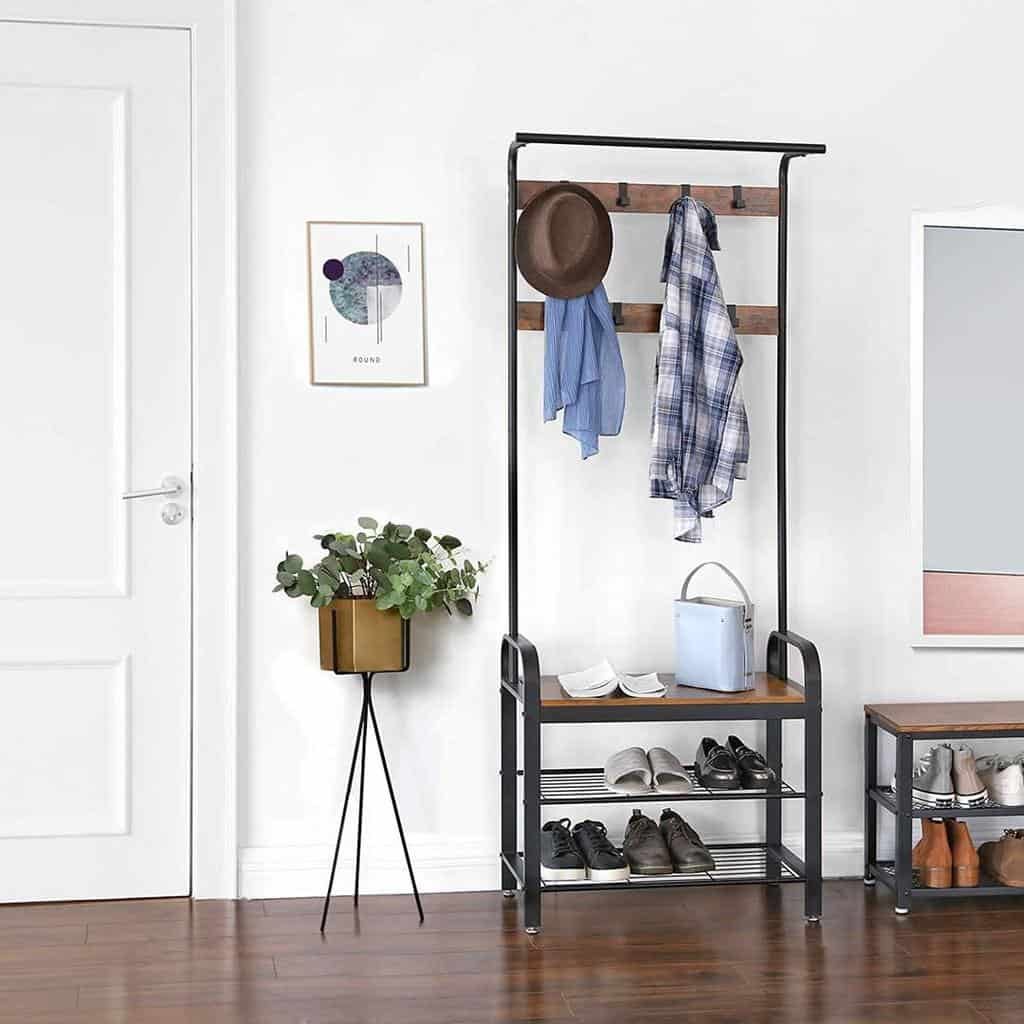 A hallway with a coat rack holding hats and clothes, a shoe rack below, a potted plant on a stand, and a wall art piece