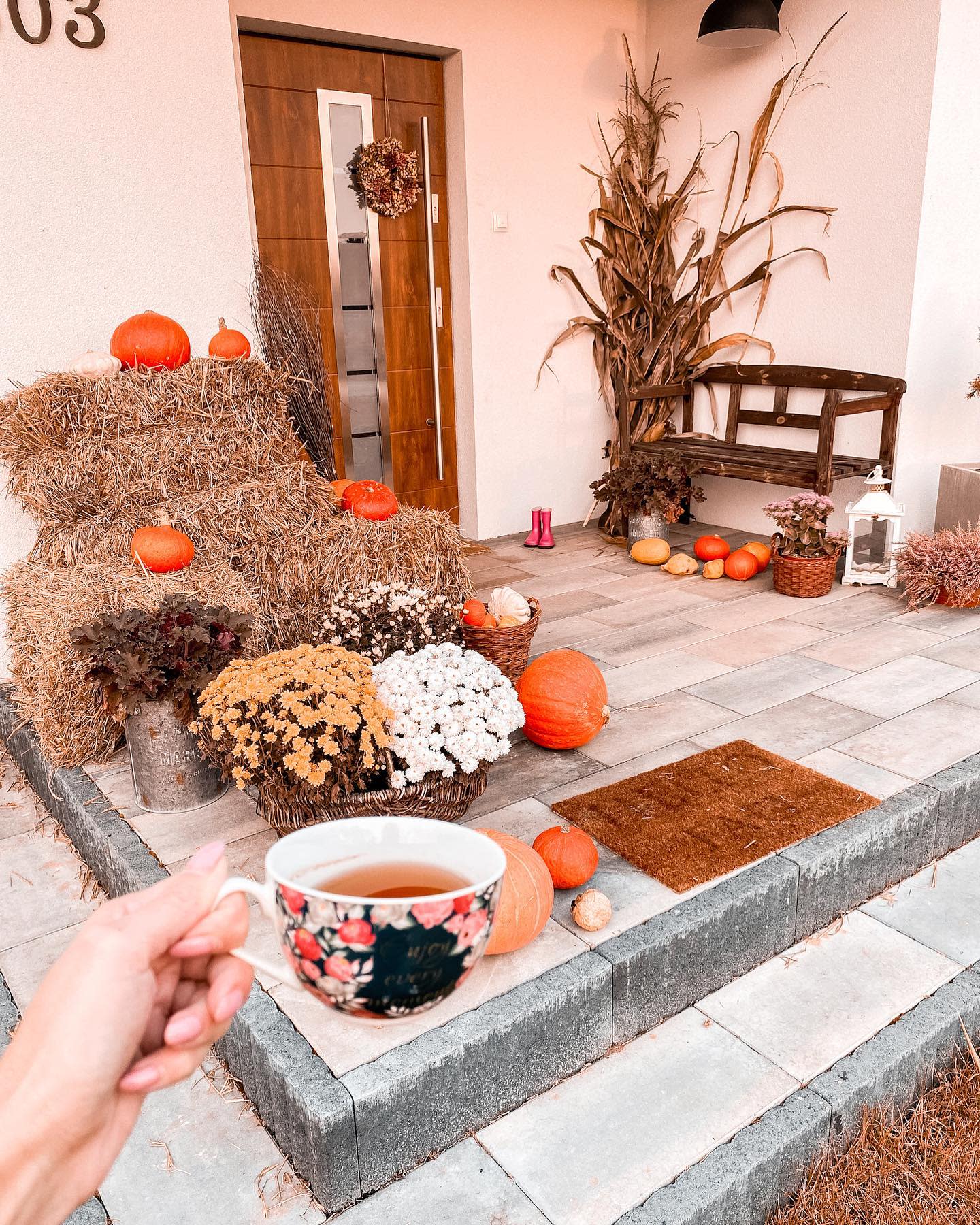 Cozy fall front porch with hay bales, pumpkins, mums, cornstalks, and a rustic bench, with a hand holding a cup of tea in the foreground