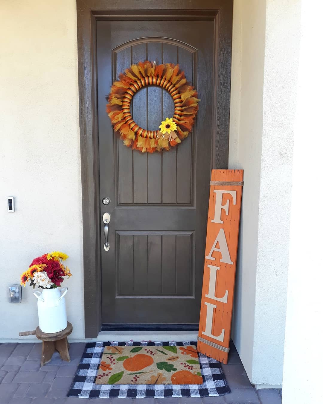 Front door with a fall wreath, "FALL" sign, bouquet of flowers in a jug, and a doormat with pumpkins on a checkered pattern