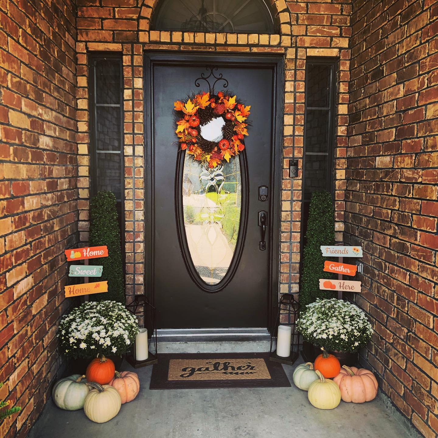 Brick porch with black oval glass door, autumn wreath, white flowers, pumpkins, and welcoming signs