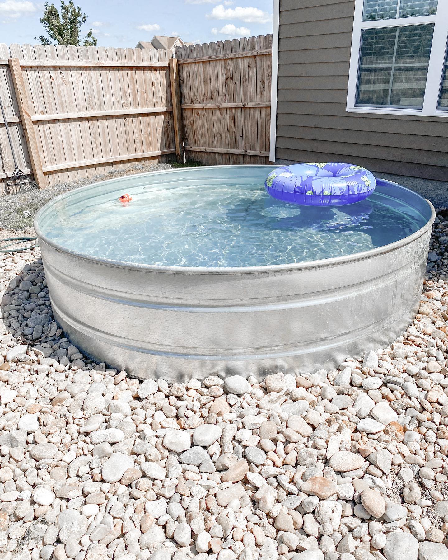 Round metal stock tank pool filled with clear water, a purple inner tube, and a toy float, surrounded by rocks near a wooden fence