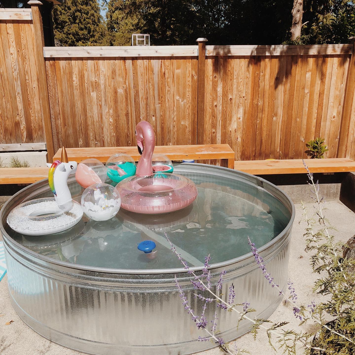 A metal tub filled with water, featuring unicorn and flamingo inflatable toys, sits outdoors by a wooden fence and benches