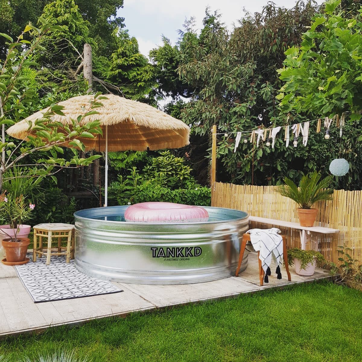 A backyard oasis with a metal tank pool, parasol, and towel on a bench, surrounded by lush plants and wooden decking